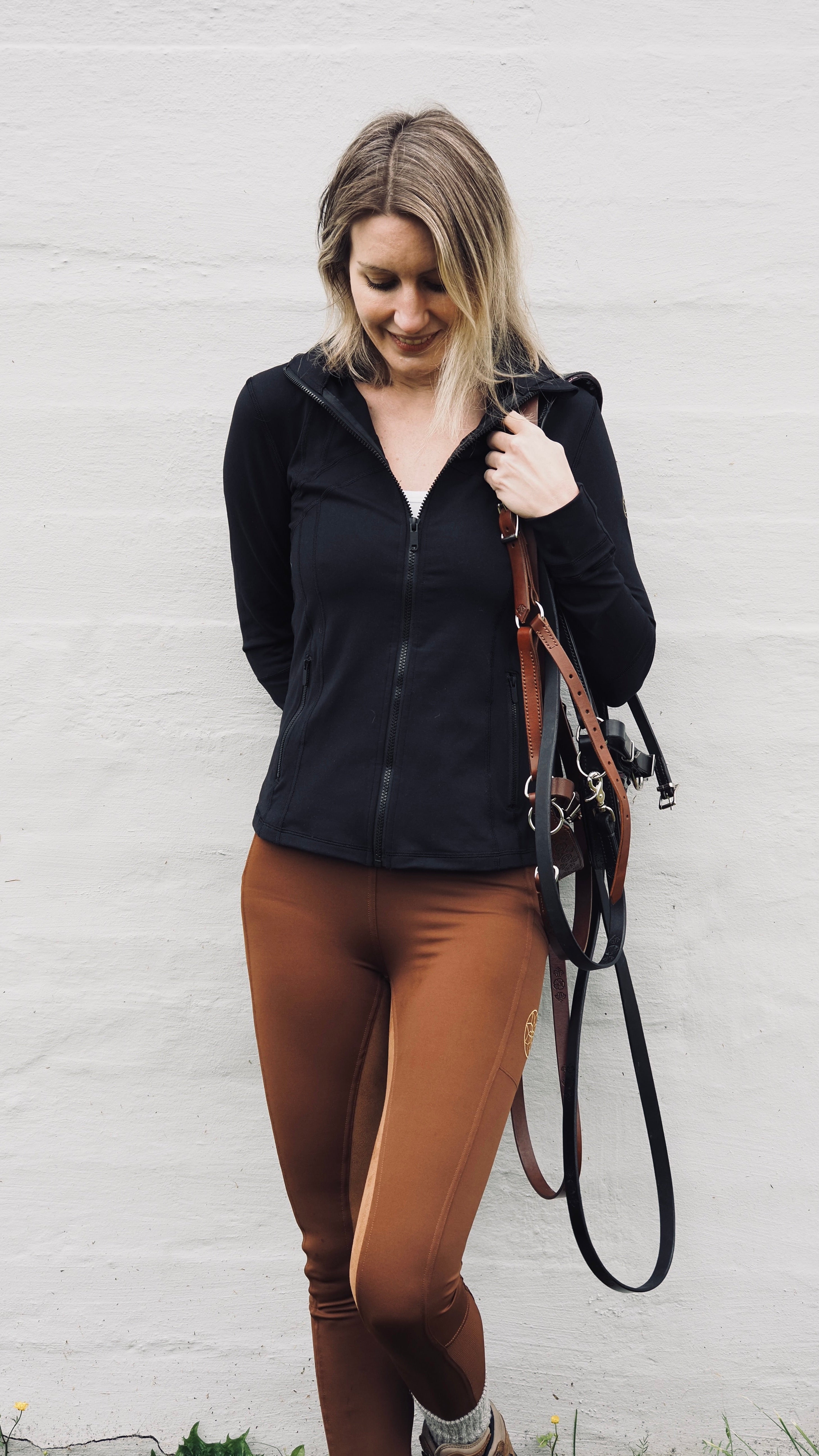 Woman in black jacket and brown riding pants holding a horse bridle against a white wall.