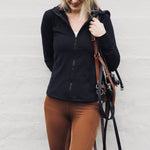 Woman in black jacket and brown riding pants holding a horse bridle against a white wall.